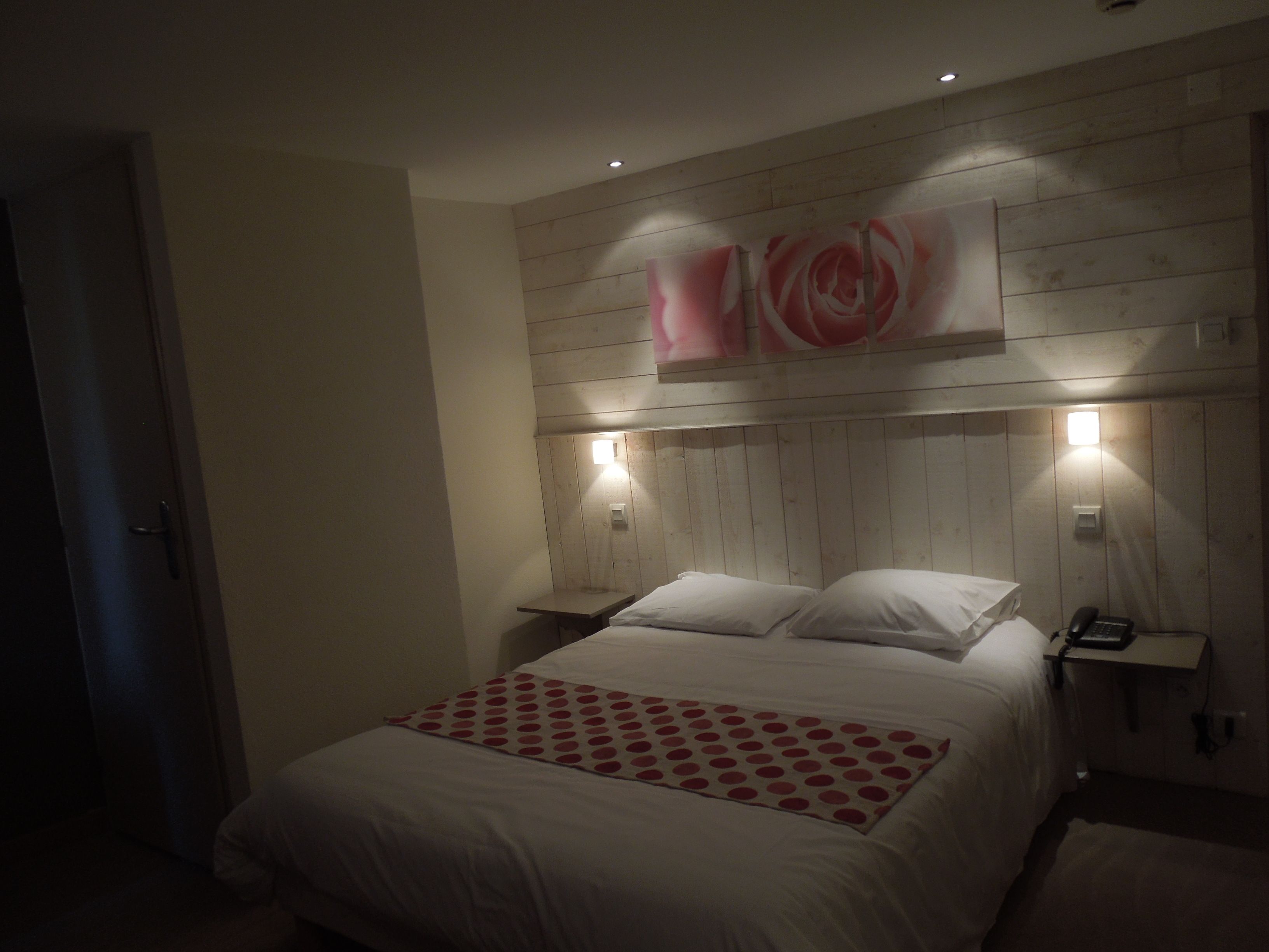 A bedroom with a wooden headboard, a bed with white bedding and a red polka dot throw, bedside lamps, and a nightstand with a phone.