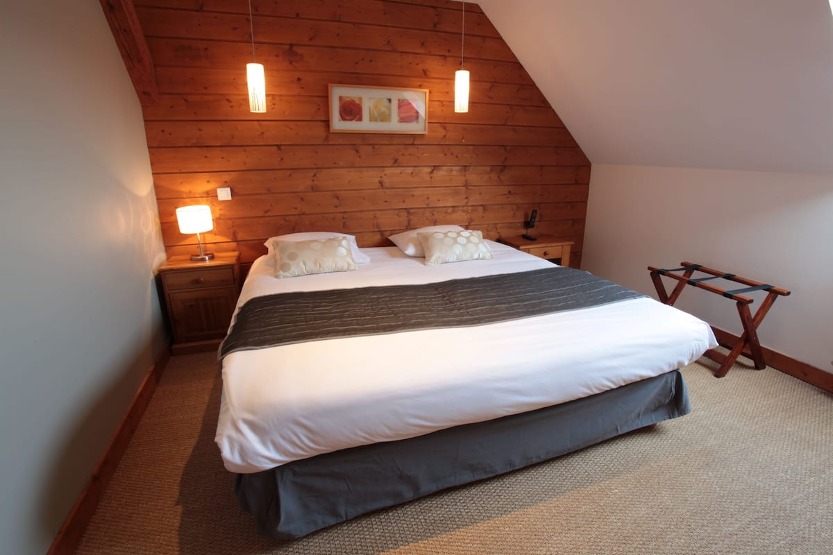 A bedroom with a sloped ceiling, wooden panel walls, a bed with white and dark bedding, two bedside lamps, and a framed picture above the bed.