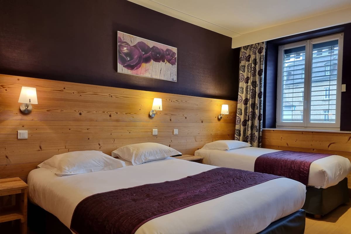 A hotel room with two beds, wooden headboard, wall-mounted lamps, a painting, and a window with curtains.