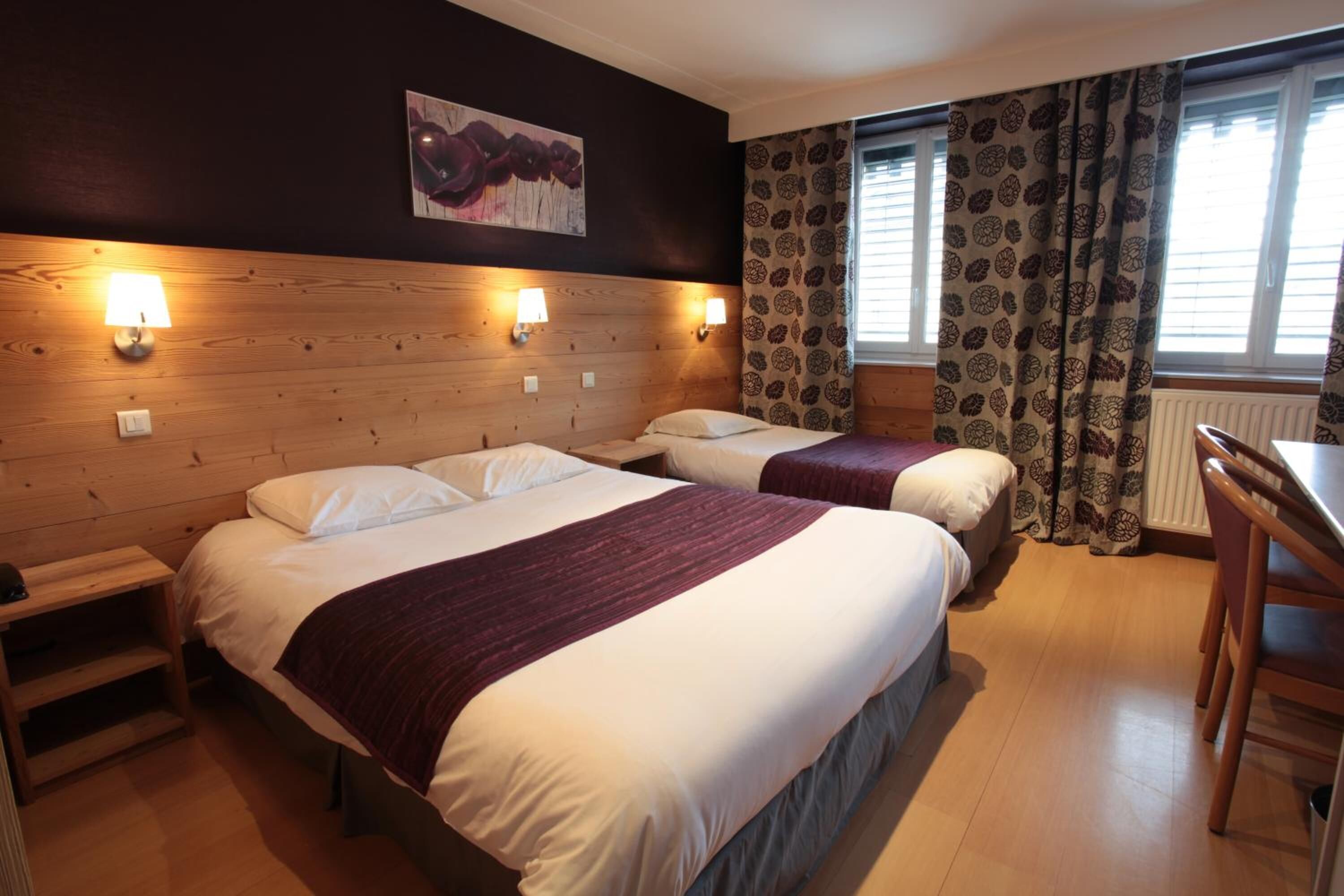 Habitación de hotel con dos camas, cabecero de madera, luces fijadas a la pared, cortinas estampadas y suelo de madera.