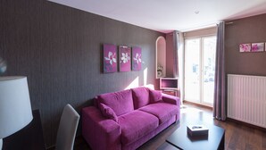 Ein modernes Wohnzimmer mit einem violetten Sofa, einem dunklen Couchtisch und einer Lampe. An den Wänden hängen drei Blumenbilder, und ein großes Fenster ist mit Vorhängen drapiert.