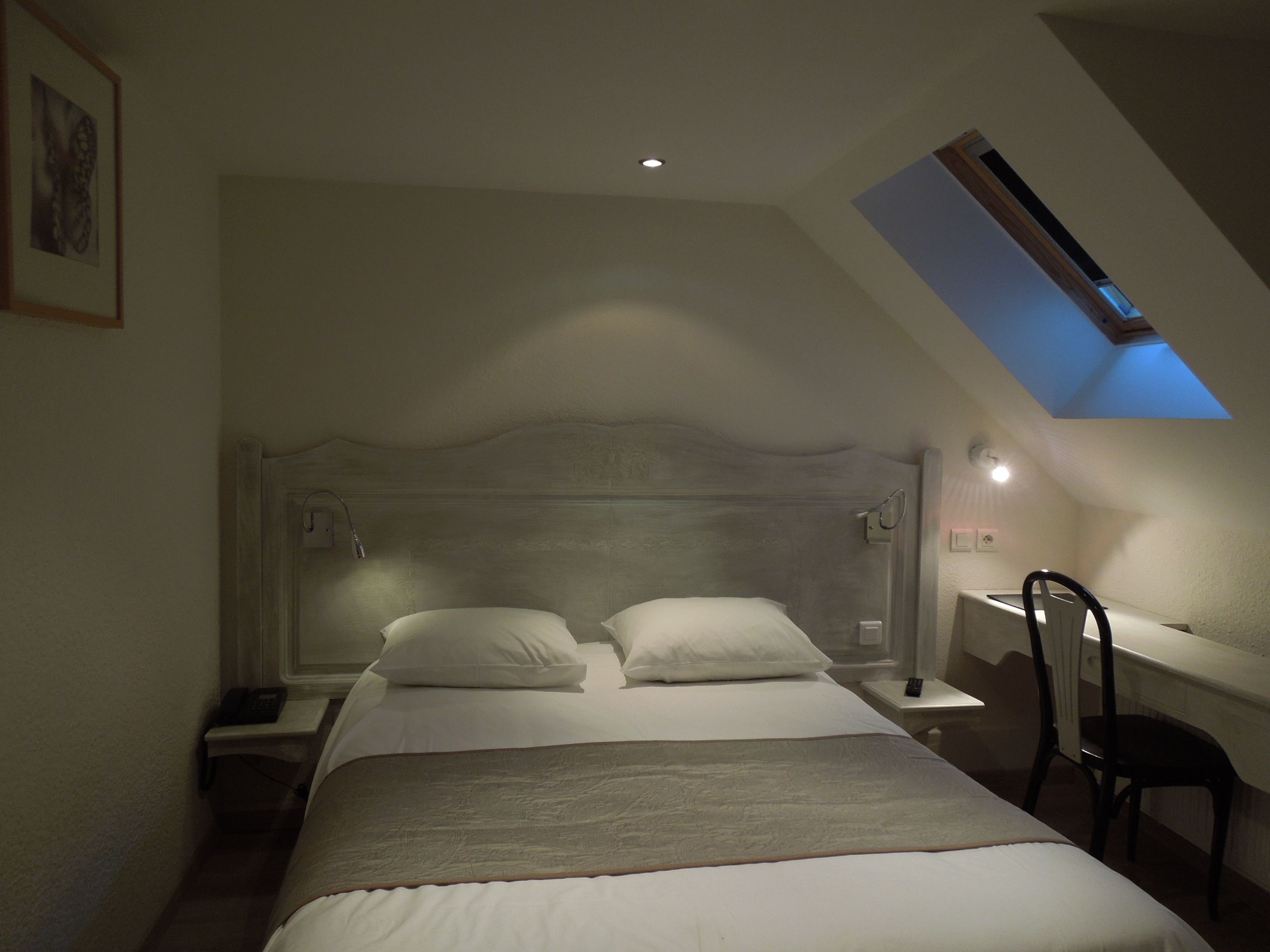 A bedroom with a sloped ceiling, a bed with white linens, a bedside table with a lamp, and a chair.