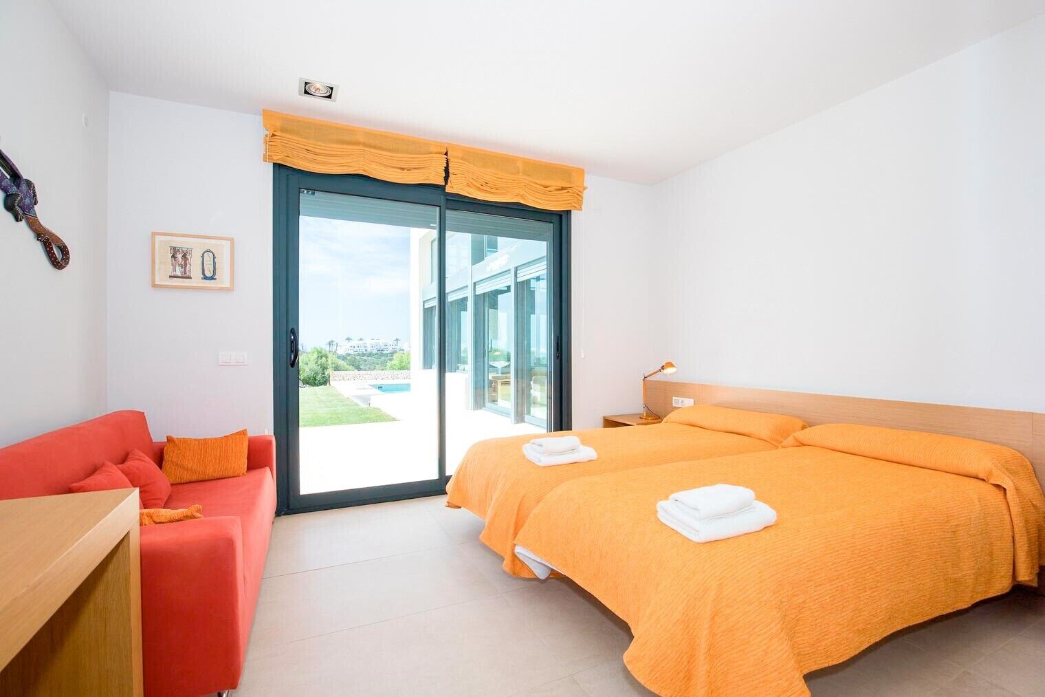A bedroom with a bed, a red sofa, a wooden nightstand, and a large window with a view of a swimming pool.