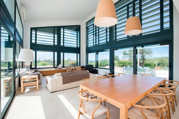 A modern living room with a wooden dining table, sofas, and a TV. Large windows offer views of the outdoors.