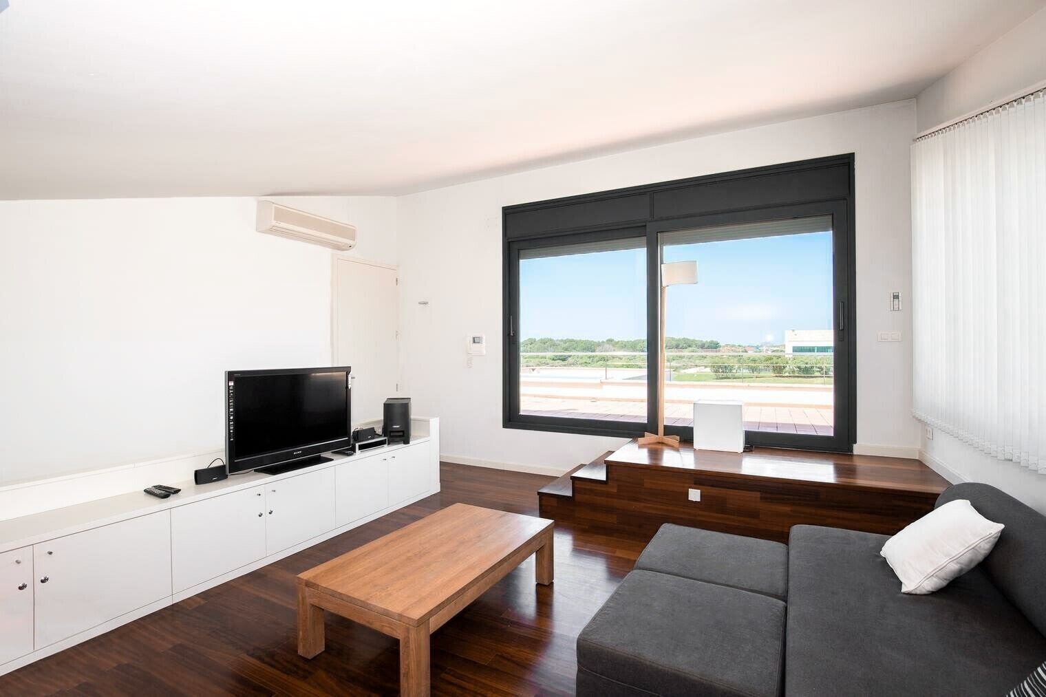 A modern living room with a flat-screen TV, wooden coffee table, grey sofa, and a large window offering a view of greenery and a building.