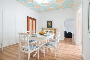 A dining area with a round table, white chairs, a fruit bowl, and a painting on the wall.
