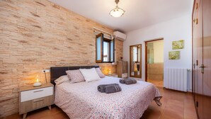 Une chambre avec un mur en pierre, un lit recouvert d’une couvre-lit à motifs floraux, une table de chevet avec une lampe et une armoire en bois.