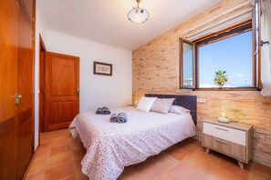 A bedroom with a wooden door, a bed with rolled towels, a nightstand with a lamp, and a window with a view.