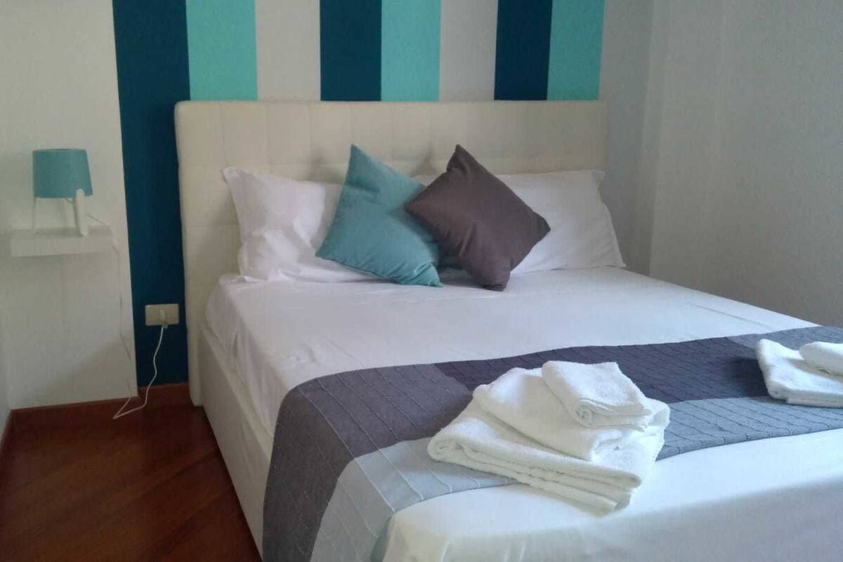 Un letto rifatto con cuscini e un asciugamano piegato, un comodino con una lampada e una parete con righe verticali.