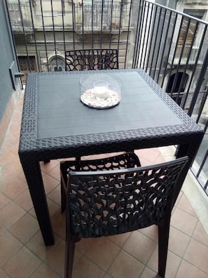 Un piccolo tavolo esterno con piano in vetro e una candela, circondato da sedie nere con decorazioni elaborate, su un balcone con ringhiera in metallo e vista su edifici sullo sfondo.