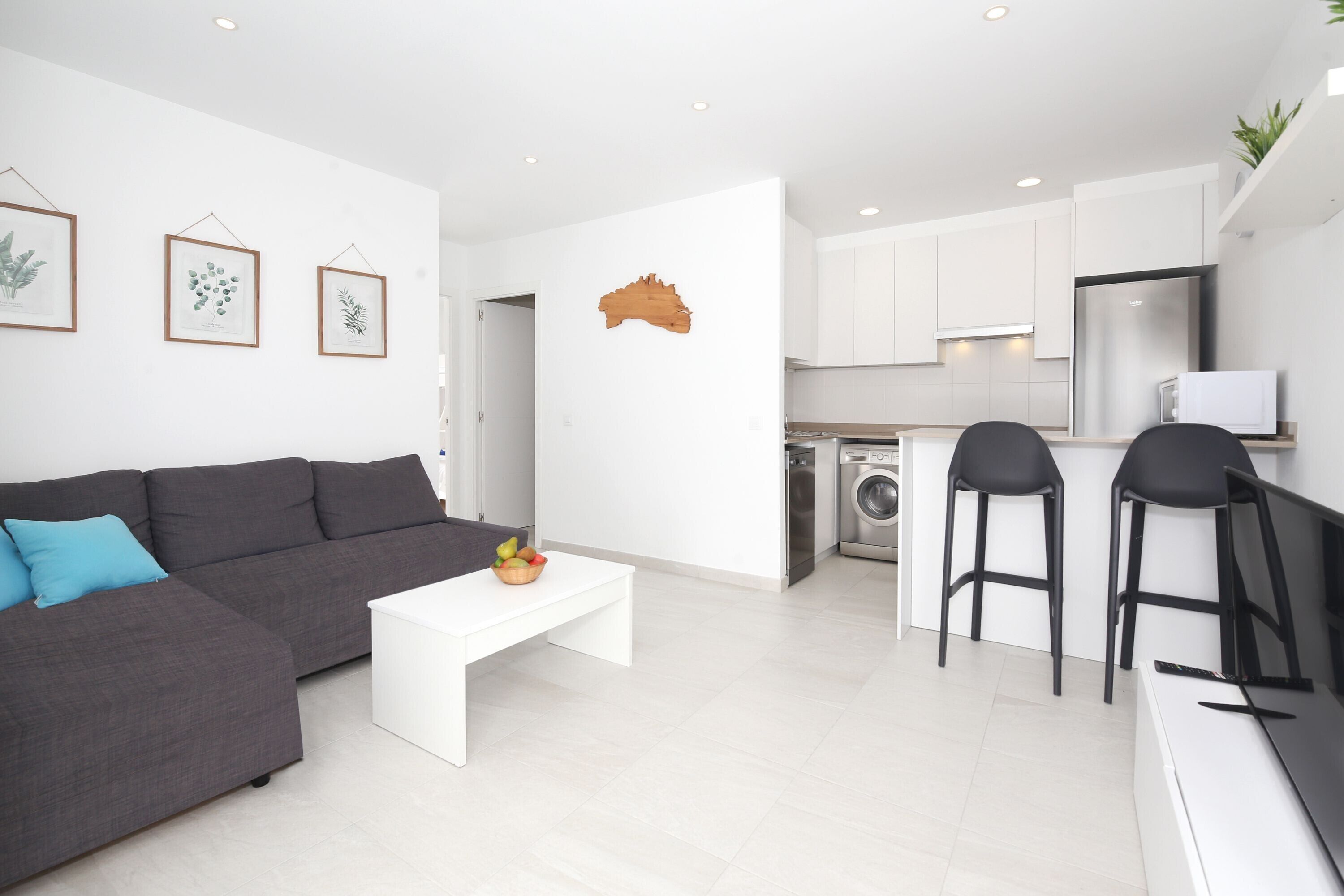 Un salon moderne avec un canapé gris, une table basse blanche et un coin cuisine équipé d’un lave-linge et de deux tabourets de bar.