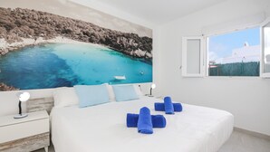 Ein Schlafzimmer mit einem großen Bett, weißer Bettwäsche, einem Nachttisch mit Lampe und einem Gemälde einer Küstenlandschaft.