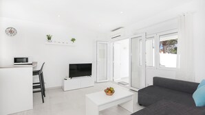 Ein modernes Wohnzimmer mit grauem Sofa, weißem Couchtisch, Flachbildfernseher, einer kleinen Küchenzeile mit Mikrowelle und einer Pflanze an der Wand.