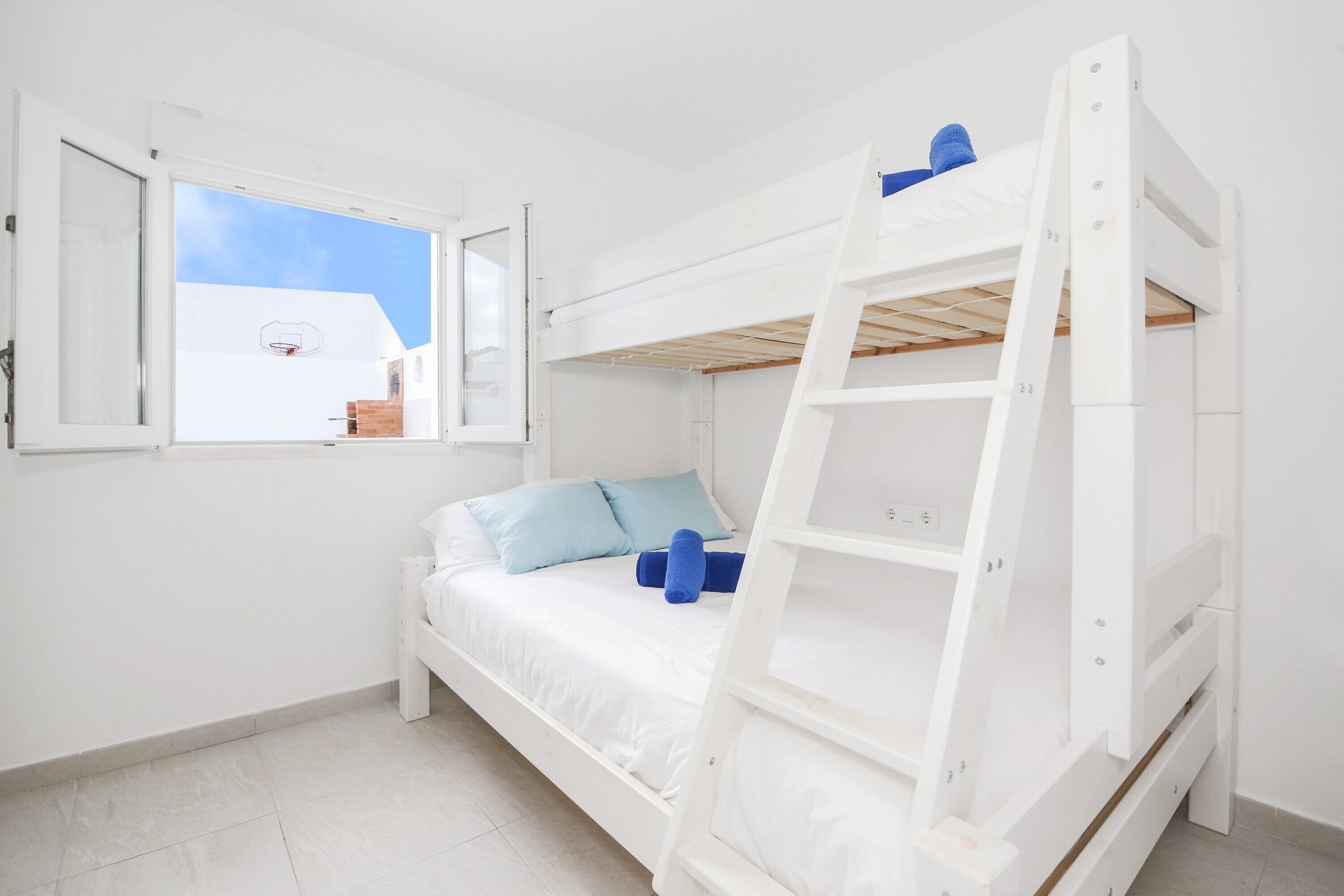 Un lit superposé blanc avec une échelle, une fenêtre aux volets ouverts et une vue sur un bâtiment à l’extérieur.