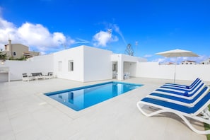 Un área de piscina exterior moderna con una piscina azul, sillas de descanso blancas y un edificio blanco al fondo.