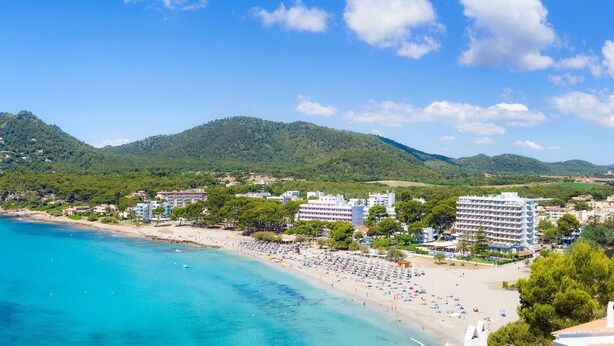 Eine Küstenlandschaft mit Strand, einem Hotel, klarem, blauem Wasser und grünen Hügeln im Hintergrund.
