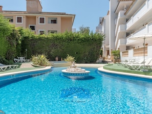 A swimming pool with lounge chairs, a fountain, and a hedge.