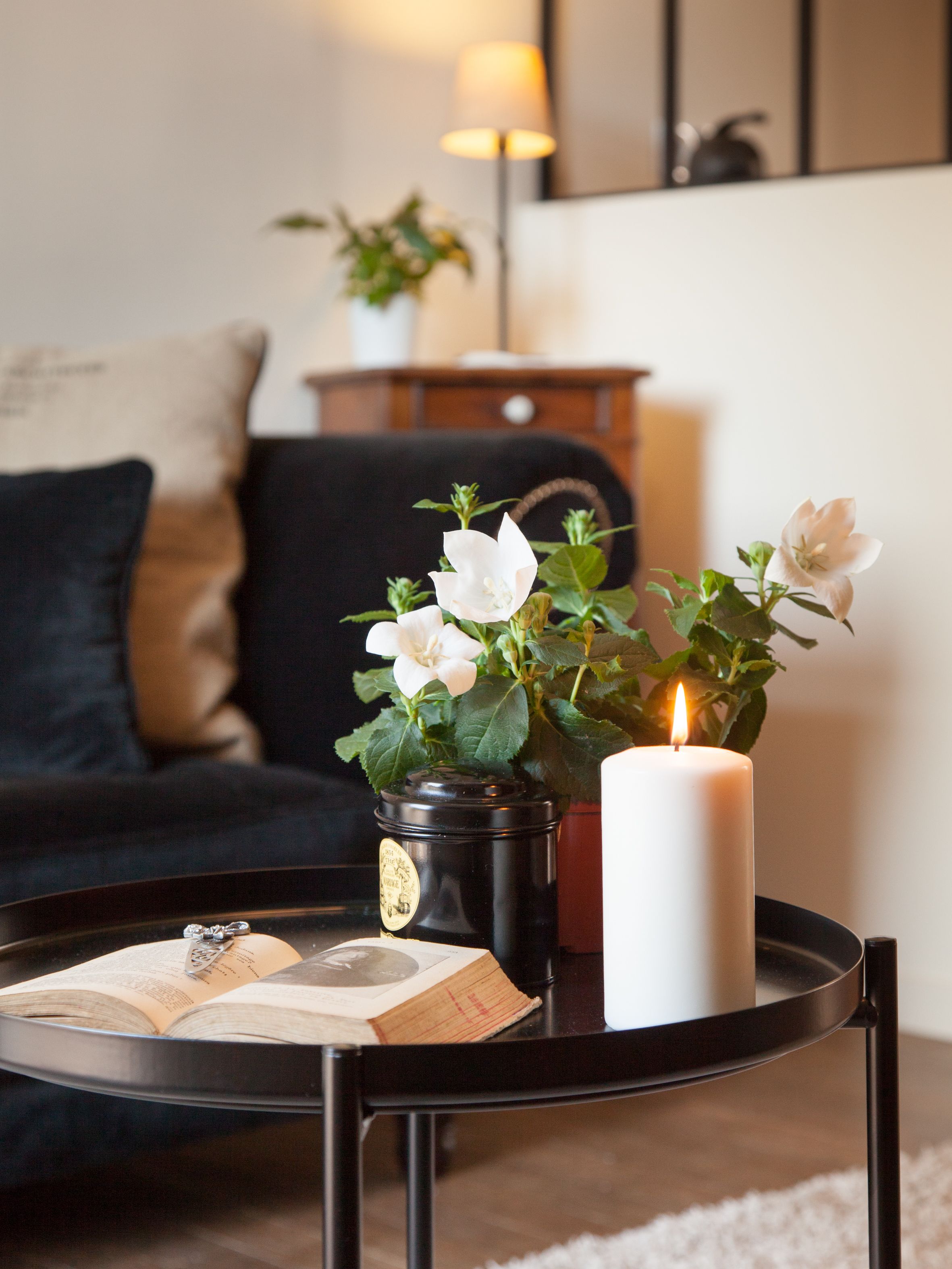 A round black table with a lit candle, a book, and a potted plant with white flowers.