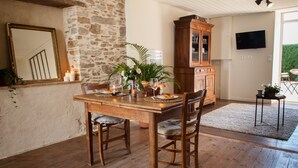 A dining area with a wooden table set for two, a stone wall, a wooden cabinet, and a television mounted on the wall. - Il etait une fois chez moi Eugénie (Le Loroux-Bottereau)