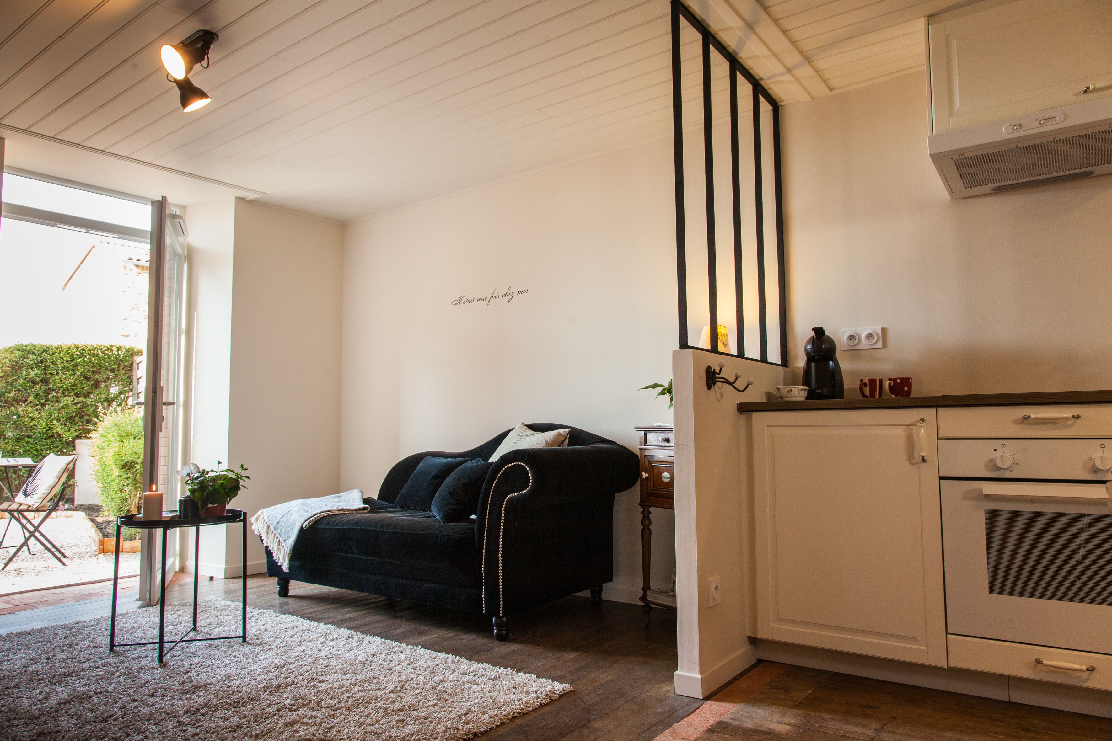 Una sala de estar acogedora con un sofá negro, una mesita con una planta y un tapete blanco. Hay una zona de cocina con armarios blancos y una estufa.