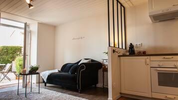 Una sala de estar acogedora con un sofá negro, una mesita con una planta y un tapete blanco. Hay una zona de cocina con armarios blancos y una estufa.