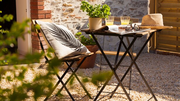 A small outdoor seating area with a table, a chair, a potted plant, a hat, and a pair of sunglasses. - Il etait une fois chez moi Eugénie (Le Loroux-Bottereau)