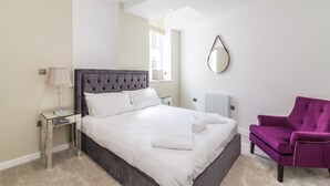 A bedroom with a large bed, a purple armchair, a bedside table with a lamp, and a wall-mounted mirror. - Hounds Gate Court (Nottingham)