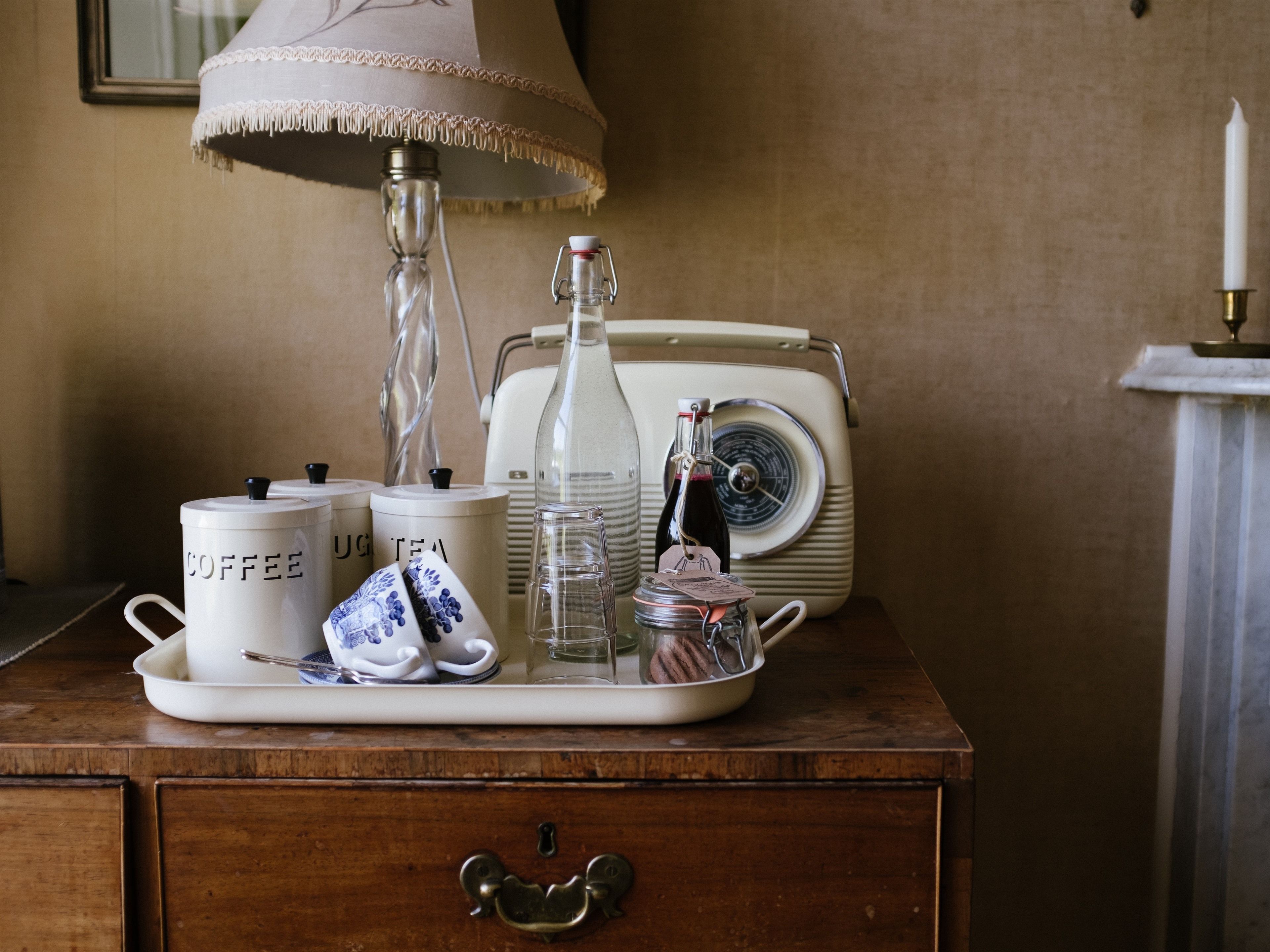 A wooden dresser with a tray holding a teapot, a bottle, a glass, and two mugs labeled "COFFEE" and "TEA". A vintage radio is in the background.
