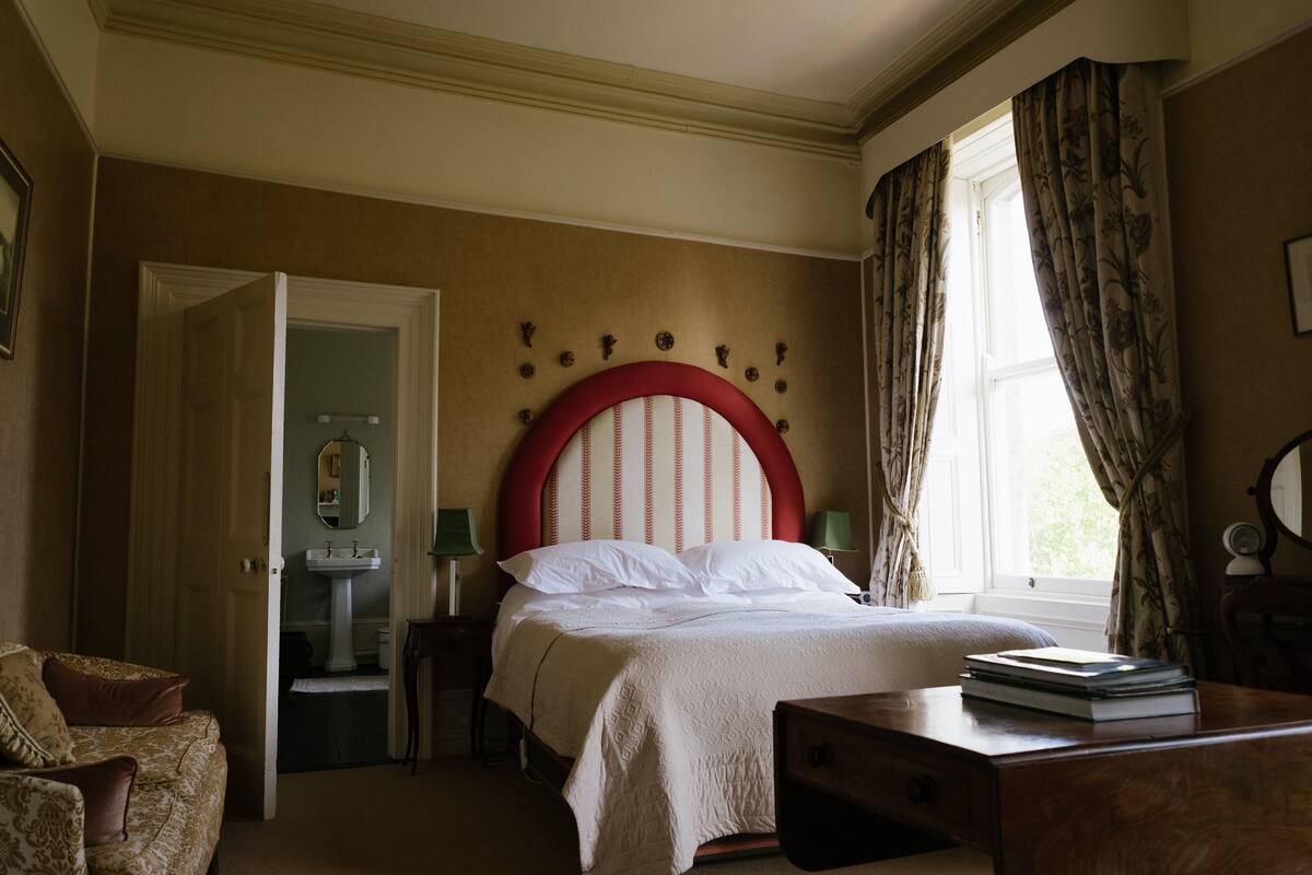 Ein Schlafzimmer mit einem großen Bett, einem Schreibtisch, einem Stuhl, einer Kommode, einem Spiegel und einem Fenster mit Vorhängen.