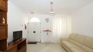 Ein Wohnzimmer mit einem Fernseher auf einem Holzständer, einem beige-farbenen Sofa, einer weißen Tür, einer Wanduhr und einer Deckenleuchte.
