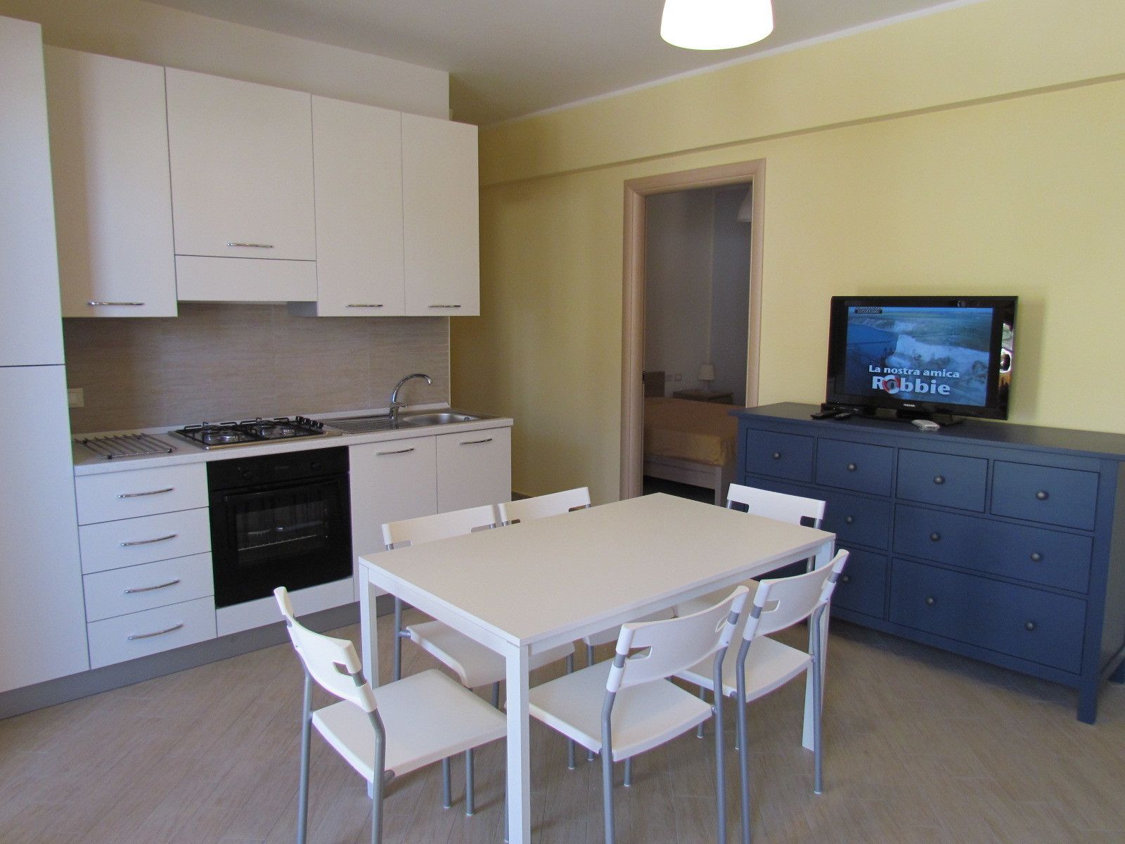 A compact kitchen with white cabinets, a stove, and a sink. There's a dining table with chairs and a TV on a blue cabinet.