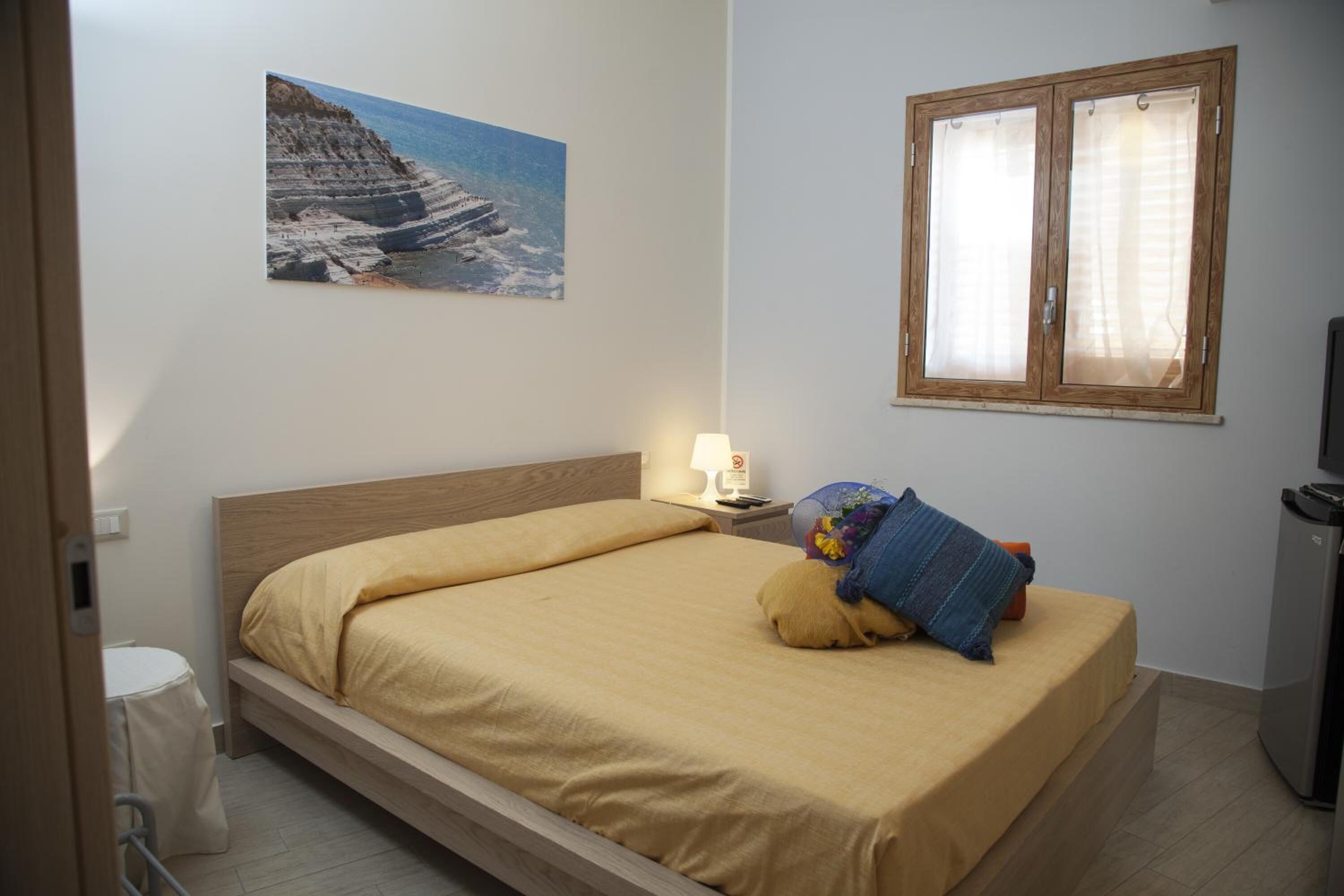 A bedroom with a bed, a nightstand, a lamp, a window, and a painting on the wall.