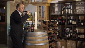 Un homme en costume, debout dans une cave, déguste du vin devant des rangées de barriques en bois et des étagères remplies de bouteilles de vin.
