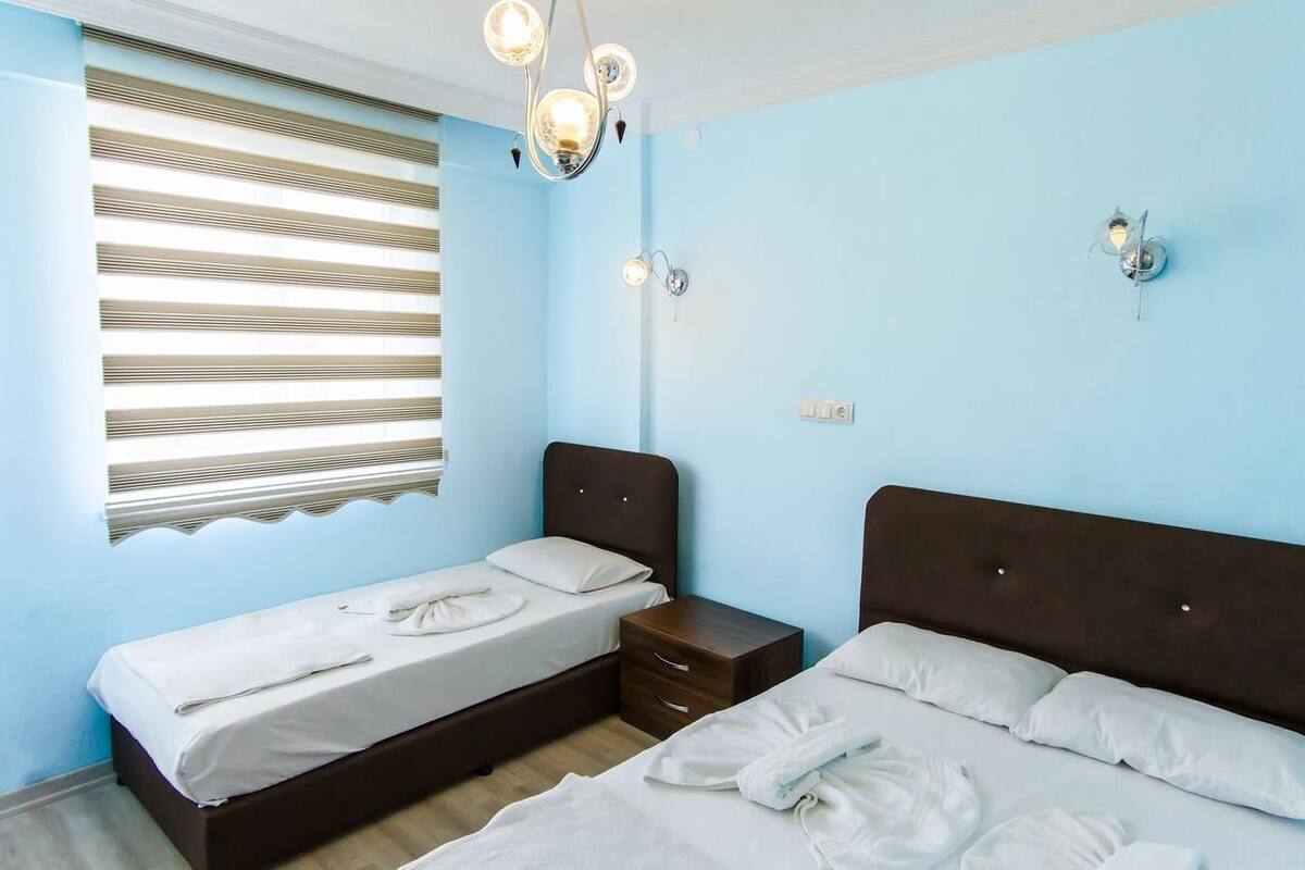 A bedroom with two beds, a window with blinds, a bedside table, and a wall-mounted light fixture.