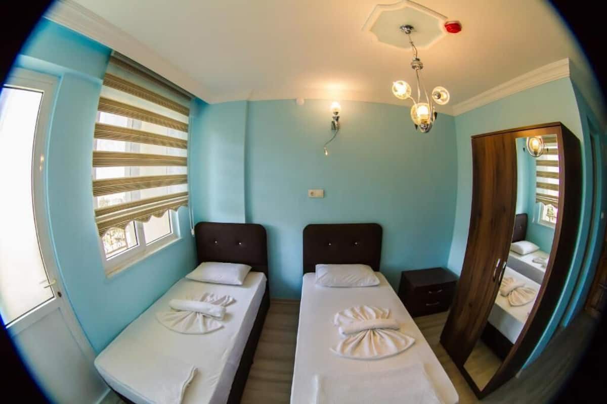 A room with two beds, a window with blinds, a wall-mounted light fixture, and a mirror.