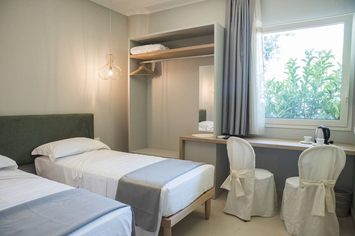 A hotel room with two beds, a desk, a chair, a hanging light, and a window with a view of greenery.