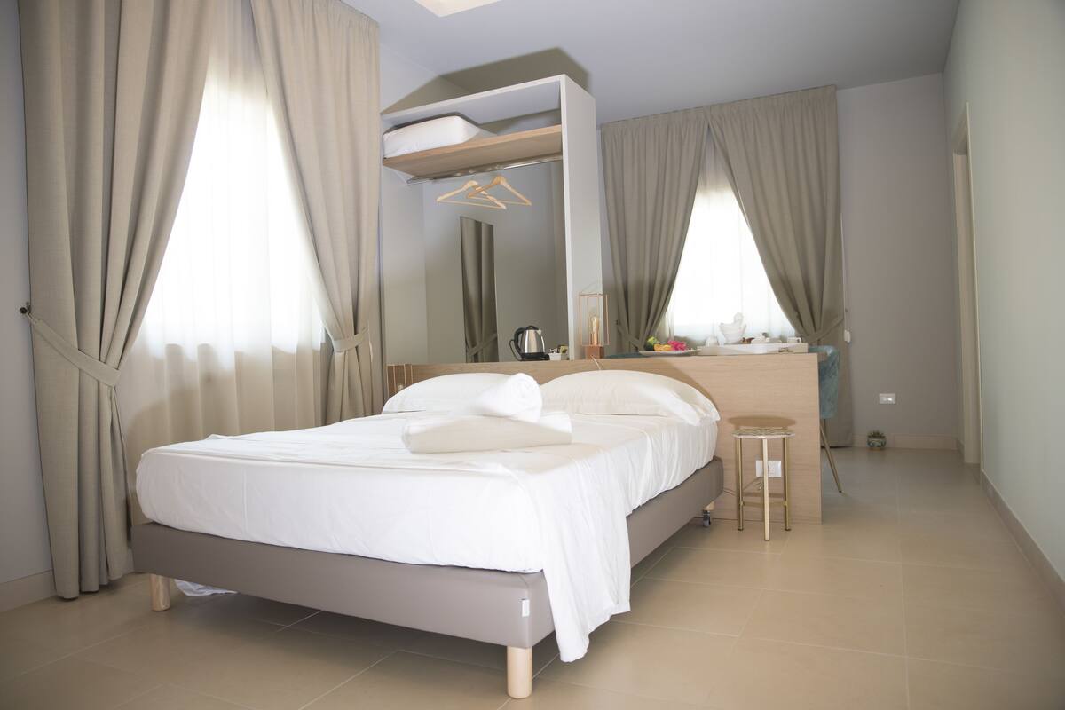 A modern bedroom with a bed, a small stool, a mirror, and a window with curtains.