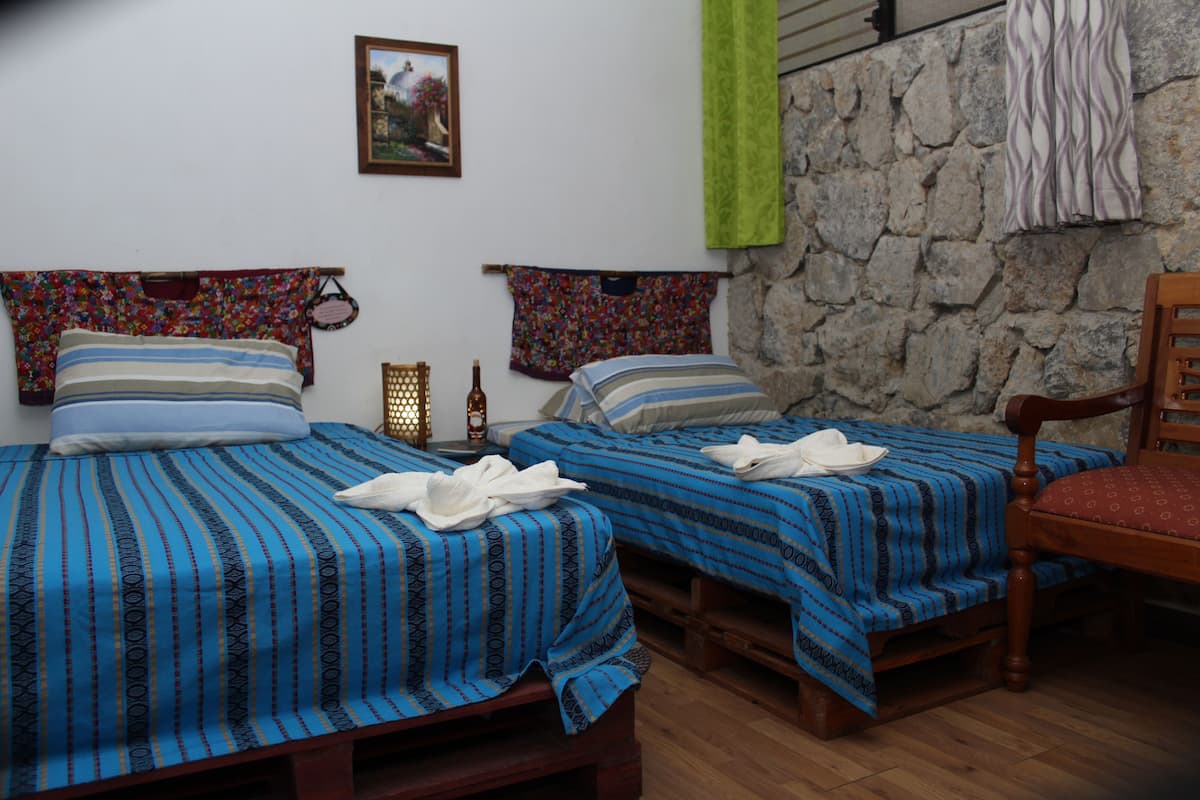 Een kamer met twee bedden, elk met een blauw gestreept laken en kussens, een houten stoel, een stenen muur en een ingelijst kunstwerk aan de muur.