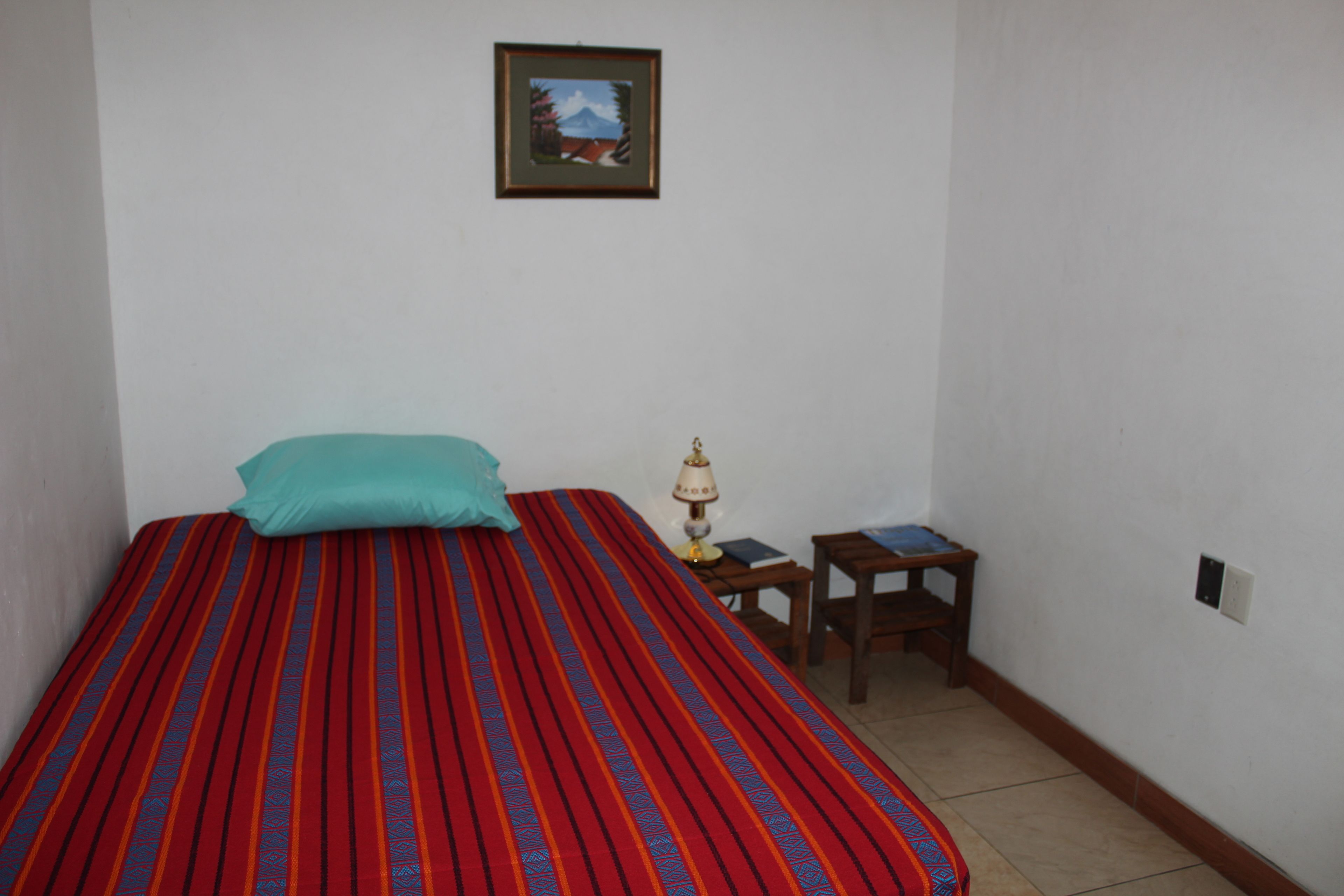 Una habitación pequeña y sencilla con una cama, una mesita de noche, una lámpara y un cuadro en la pared.