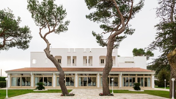 Un bâtiment moderne doté d’un toit en tuiles, de grandes fenêtres et d’une cour pavée où se trouvent deux arbres imposants.