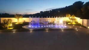 Une fontaine dotée de plusieurs jets d’eau et illuminée de lumières bleues, entourée d’une zone pavée avec des arbres et un bâtiment en arrière-plan.