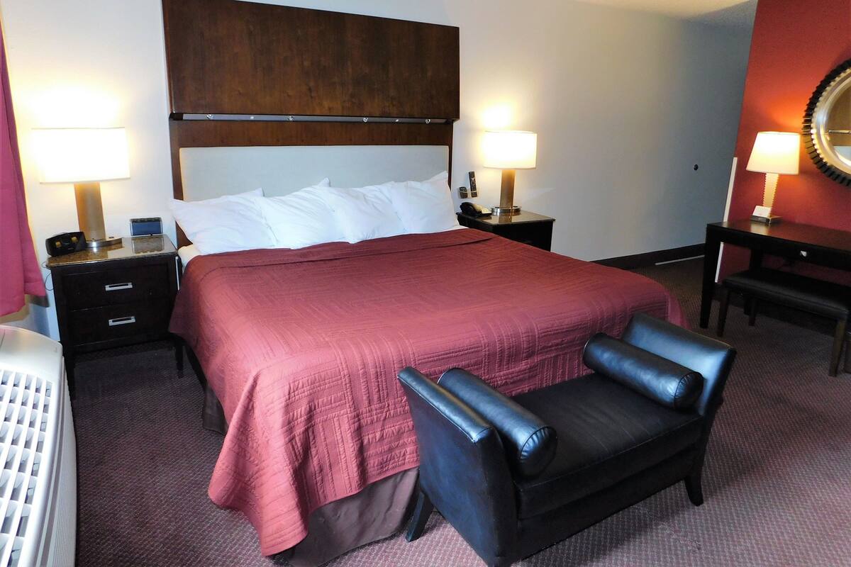 A hotel room with a large bed, two bedside tables with lamps, a wooden headboard, a black leather chair, and a desk with a mirror.