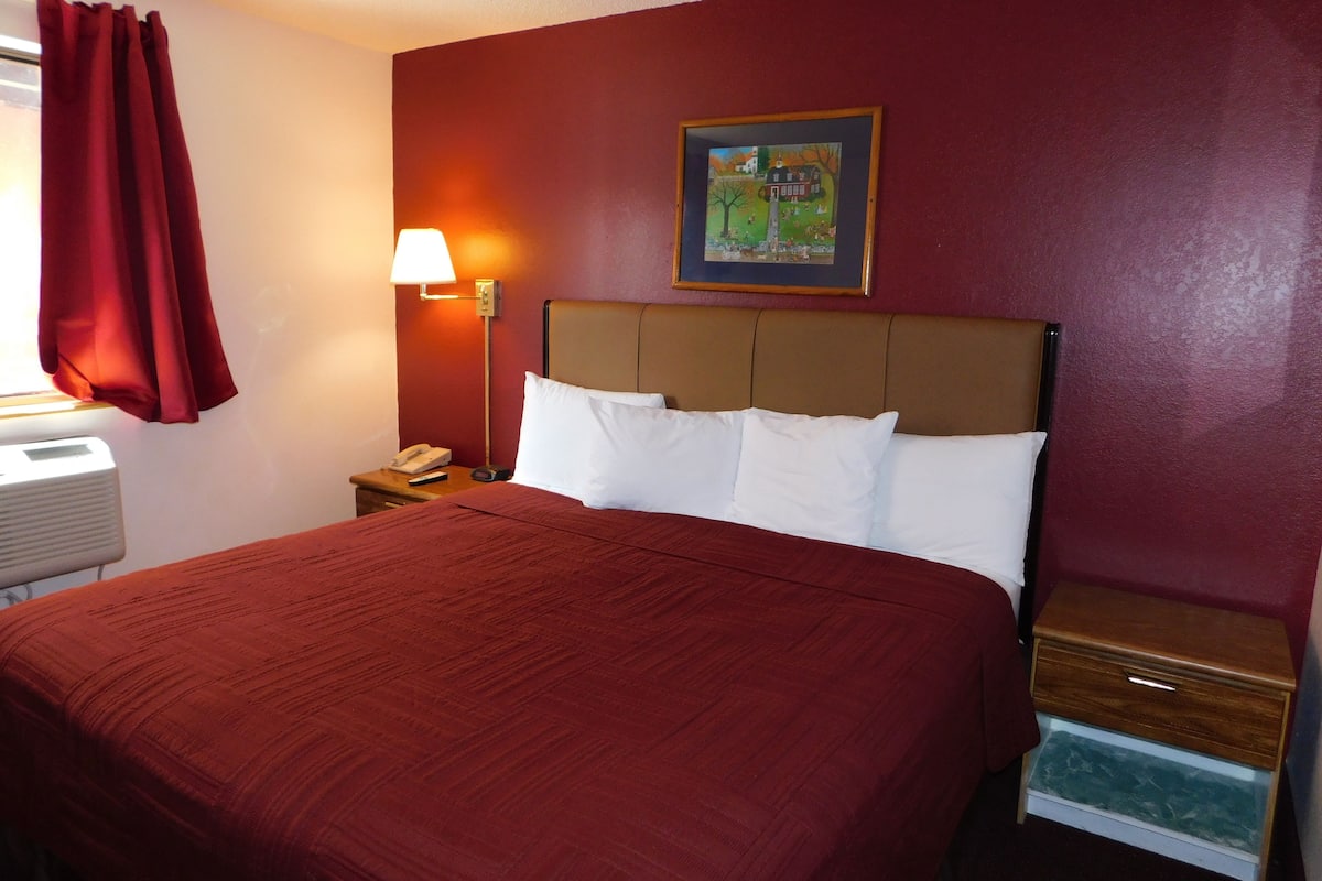A hotel room with a large bed, a nightstand with a lamp, a telephone, and a small drawer.