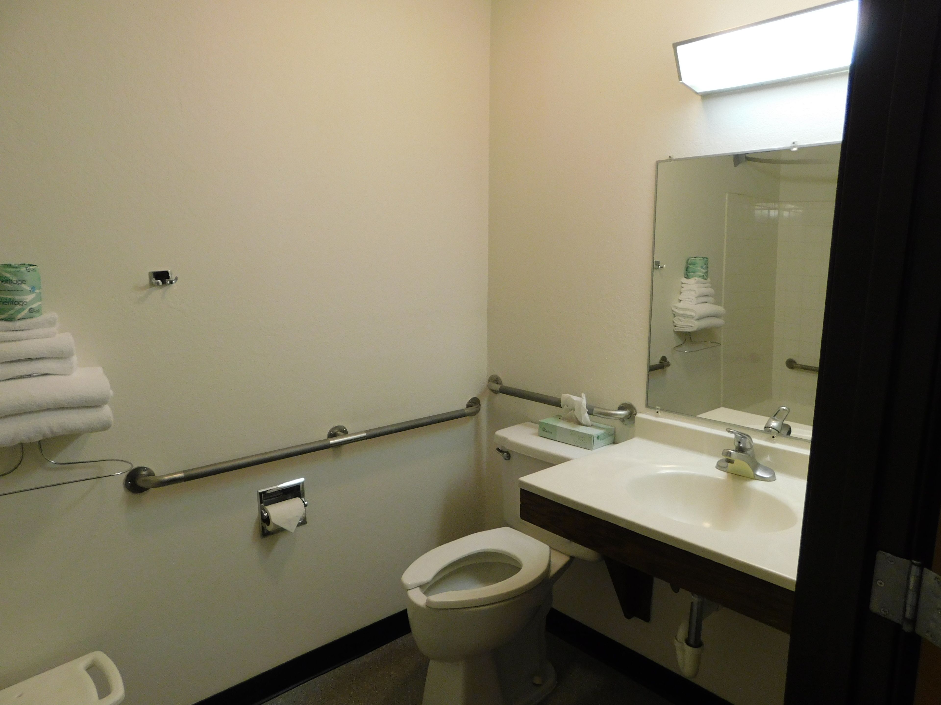 Baño estándar de hotel con inodoro, lavamanos, espejo y barras de apoyo.
