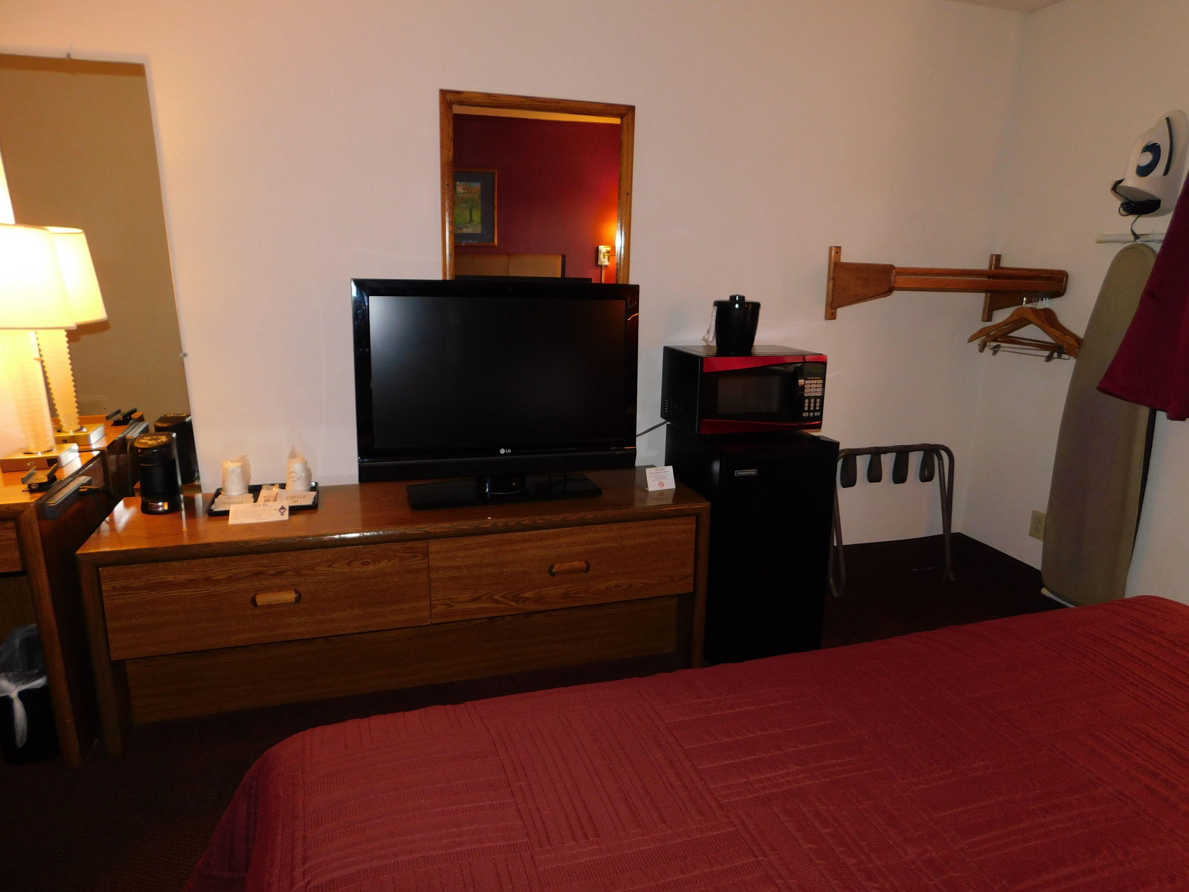 Ein Hotelzimmer mit einem Bett, einer Holzkommode mit Fernseher und Mikrowelle darauf, einem Schreibtisch mit Lampe sowie einem Kleiderständer mit Mantel.