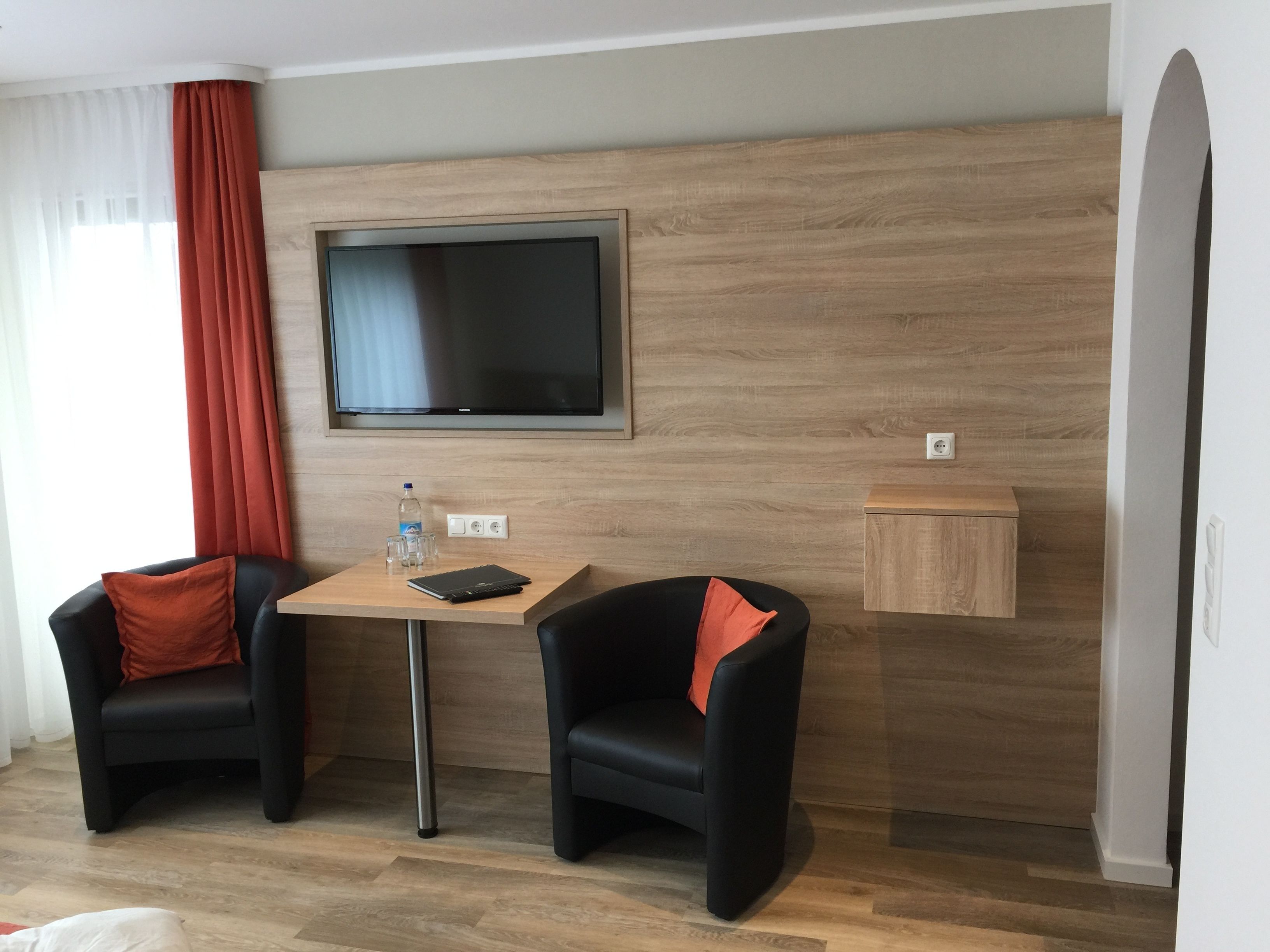 A modern hotel room with a wooden wall, a flat-screen TV, two black leather chairs with red cushions, a small wooden side table, and a water bottle on the table.