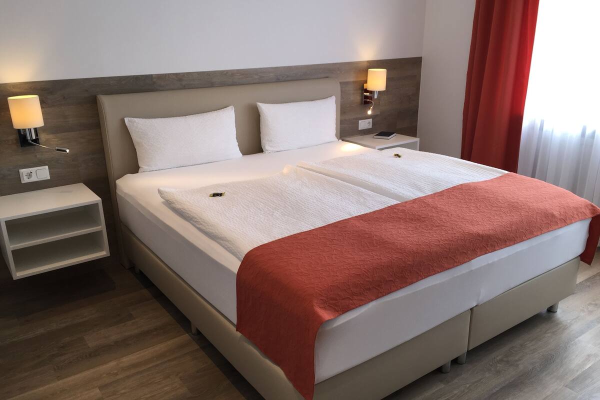 Una cama doble con una manta blanca y roja, dos almohadas blancas, un cabecero de madera, mesitas de noche con lámparas y una cortina roja.
