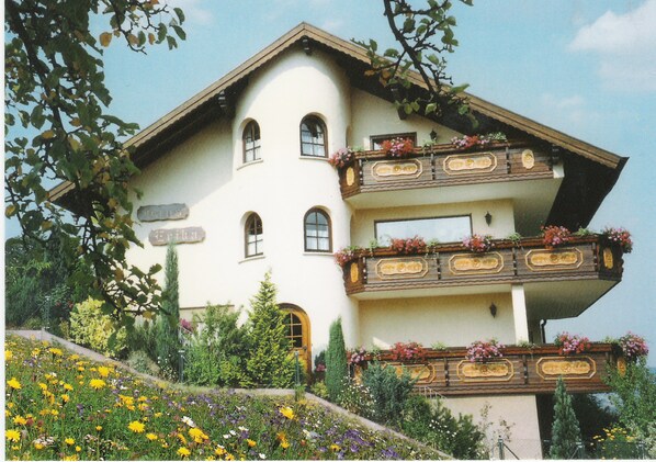 Um edifício de dois andares com varandas decoradas com flores, rodeado por um jardim com diversas plantas e flores.