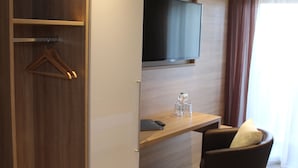 A modern hotel room with a wooden wardrobe, a flat-screen TV mounted on the wall, a desk with a chair, and a window with curtains. - Pension Haus Erika (Stadtprozelten)