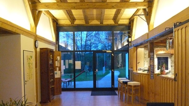 A spacious hallway with wooden beams on the ceiling, a glass door leading outside, and a small seating area with stools. - Camping Schwabenmühle (Weikersheim)